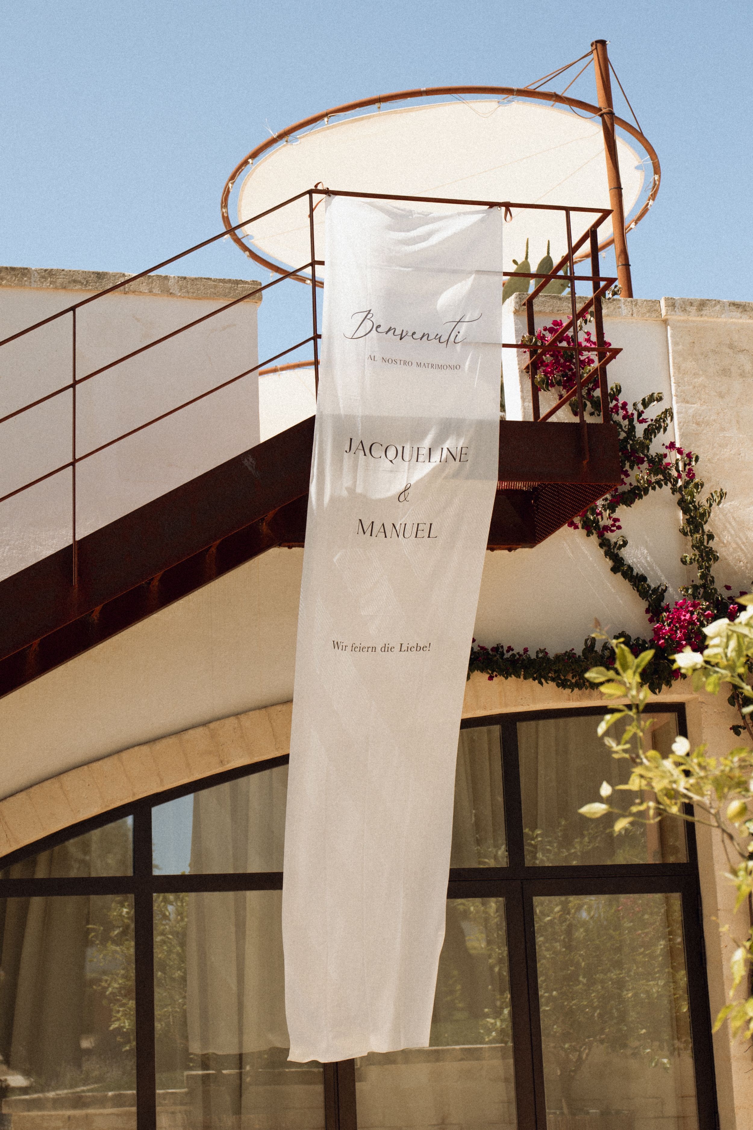 White wedding banner for Jacqueline and Manuel displayed on a Mediterranean building facade with pink flowers.