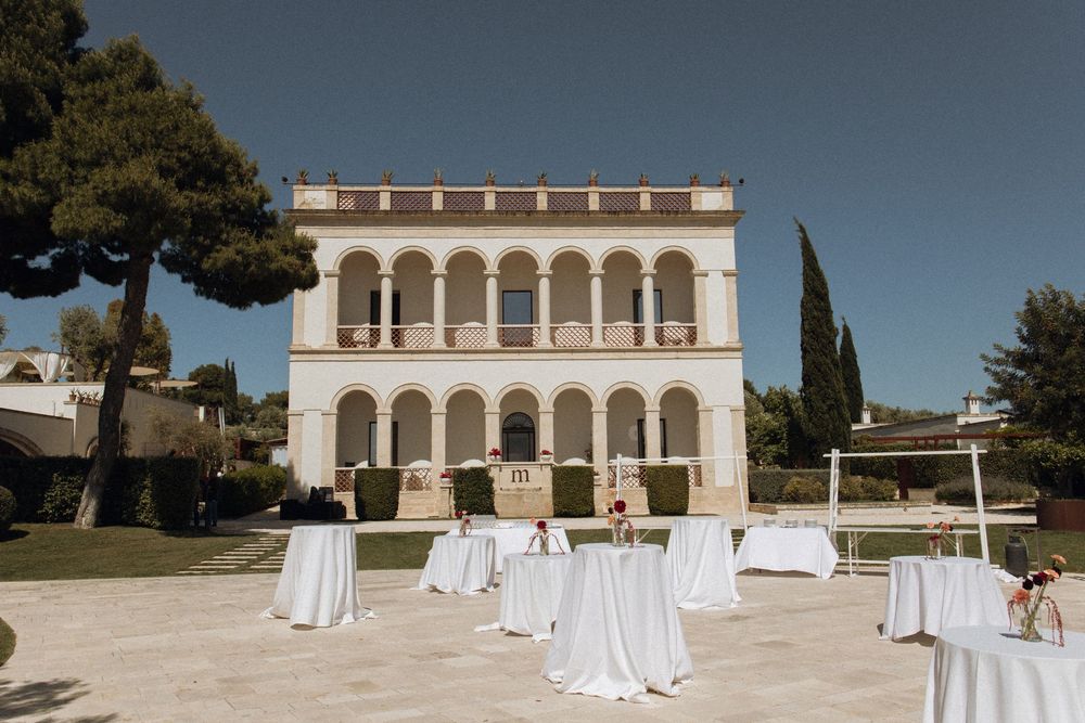 Elegant villa wedding venue with white draped cocktail tables on a sunny terrace surrounded by lush greenery.