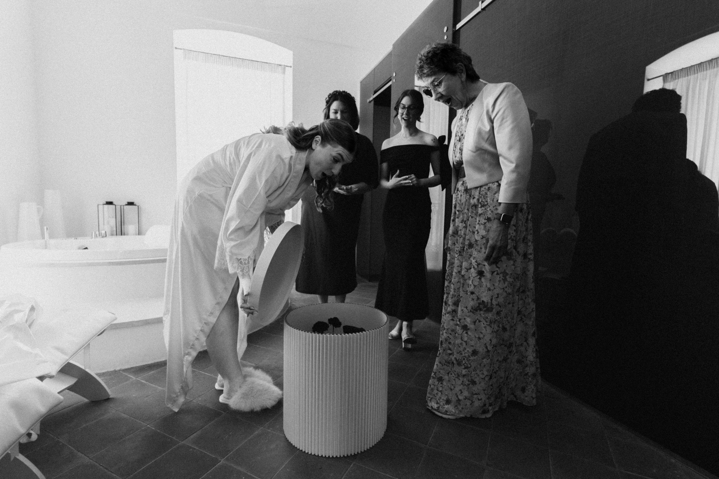Black and white photo of a bride in white dress with bridesmaids gathered around a pedestal in a bright room.