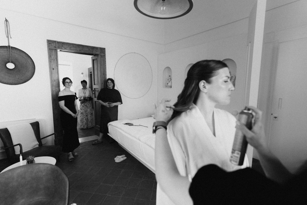 Black and white photo of a bride in a white robe getting her hair done while guests watch in the background.