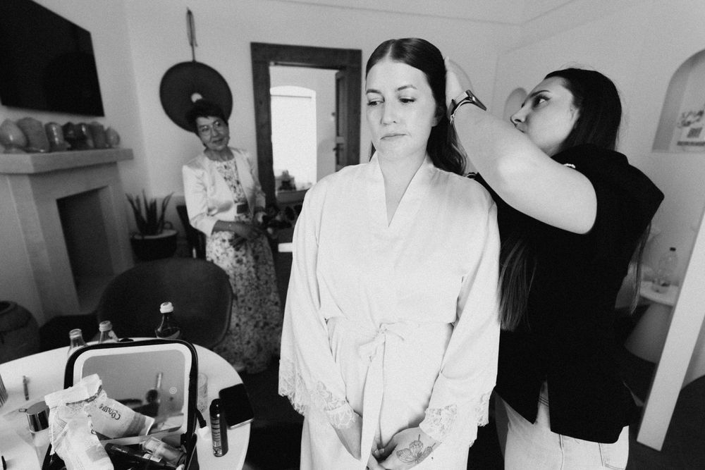 Black and white photo of bride in robe getting hair done by stylist, with guest watching in background.