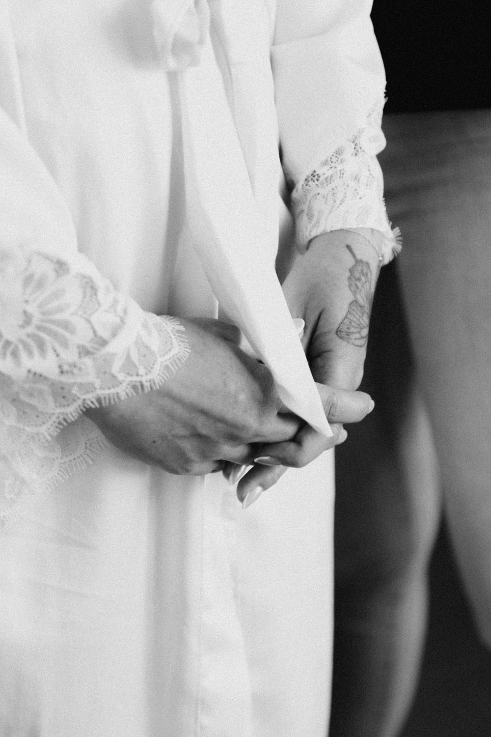 Black and white photo of bride and groom holding hands, showcasing lace wedding dress sleeve and tattoo.