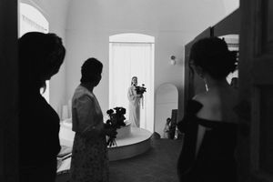 Black and white wedding photo of bride in doorway with silhouetted guests holding roses in elegant venue.