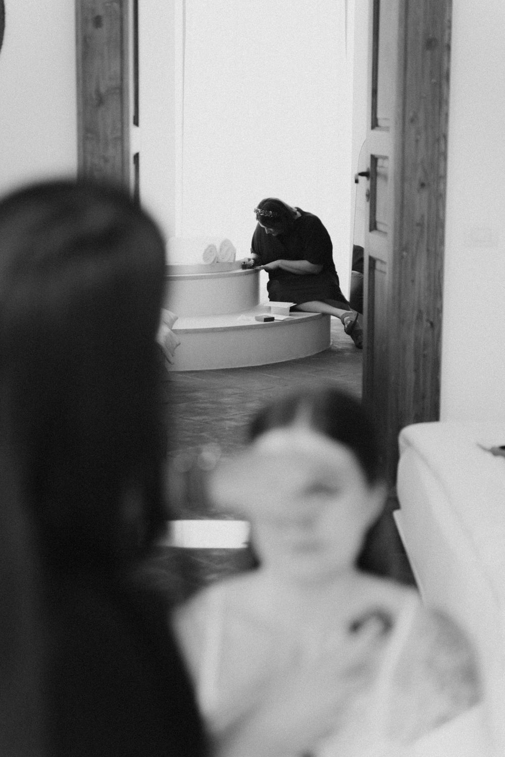 Black and white photo of woman and blurred child in foreground, with person sitting by bathtub in background.