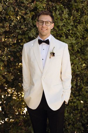 Smiling groom in cream tuxedo jacket, black bow tie, boutonniere, and glasses, posing outdoors against lush green foliage.