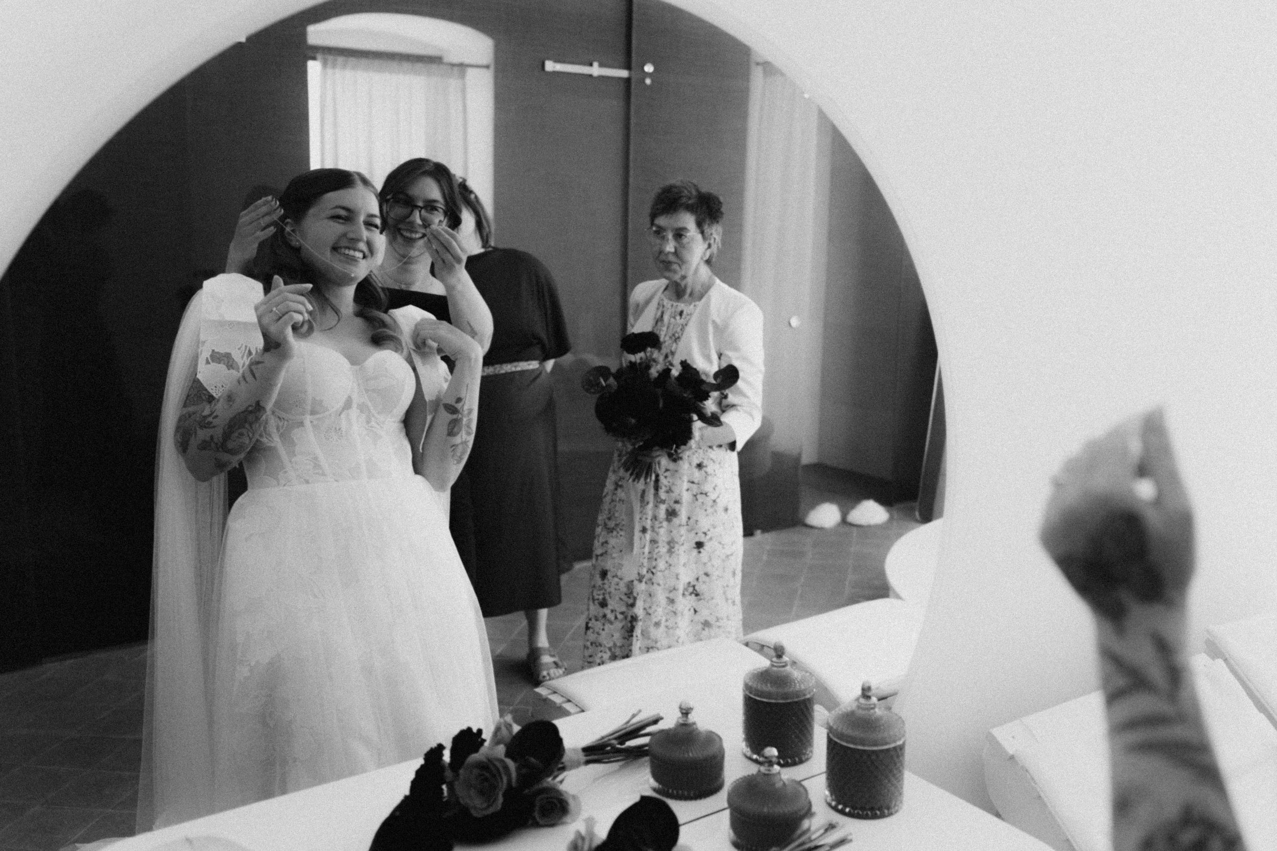 Black and white photo of a bride getting ready, adjusting her earrings in a mirror with two women assisting her.