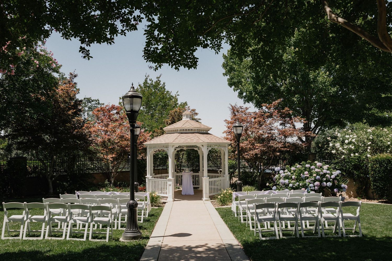 An outdoor wedding ceremony setup with white chairs flanking an aisle leading to an elegant gazebo.