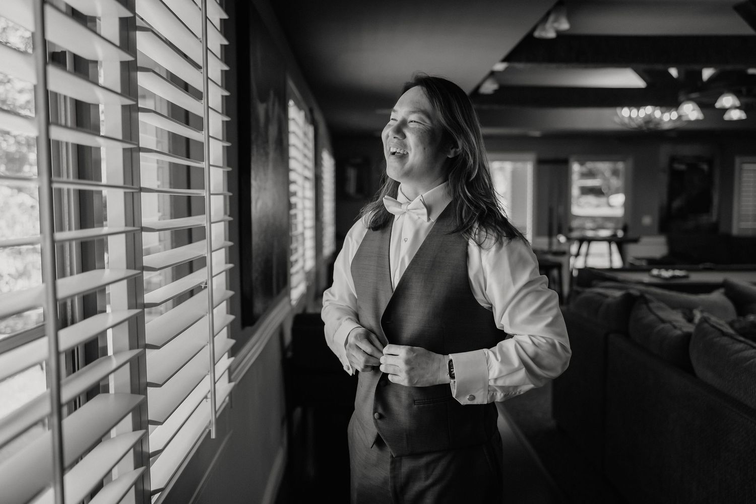 Groom adjusts his vest while standing by window blinds in a black and white wedding preparation photo.
