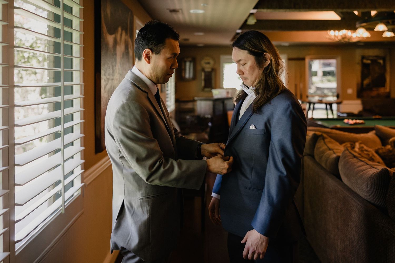 Groomsman helps partner adjust suit jacket during intimate indoor wedding day preparations.