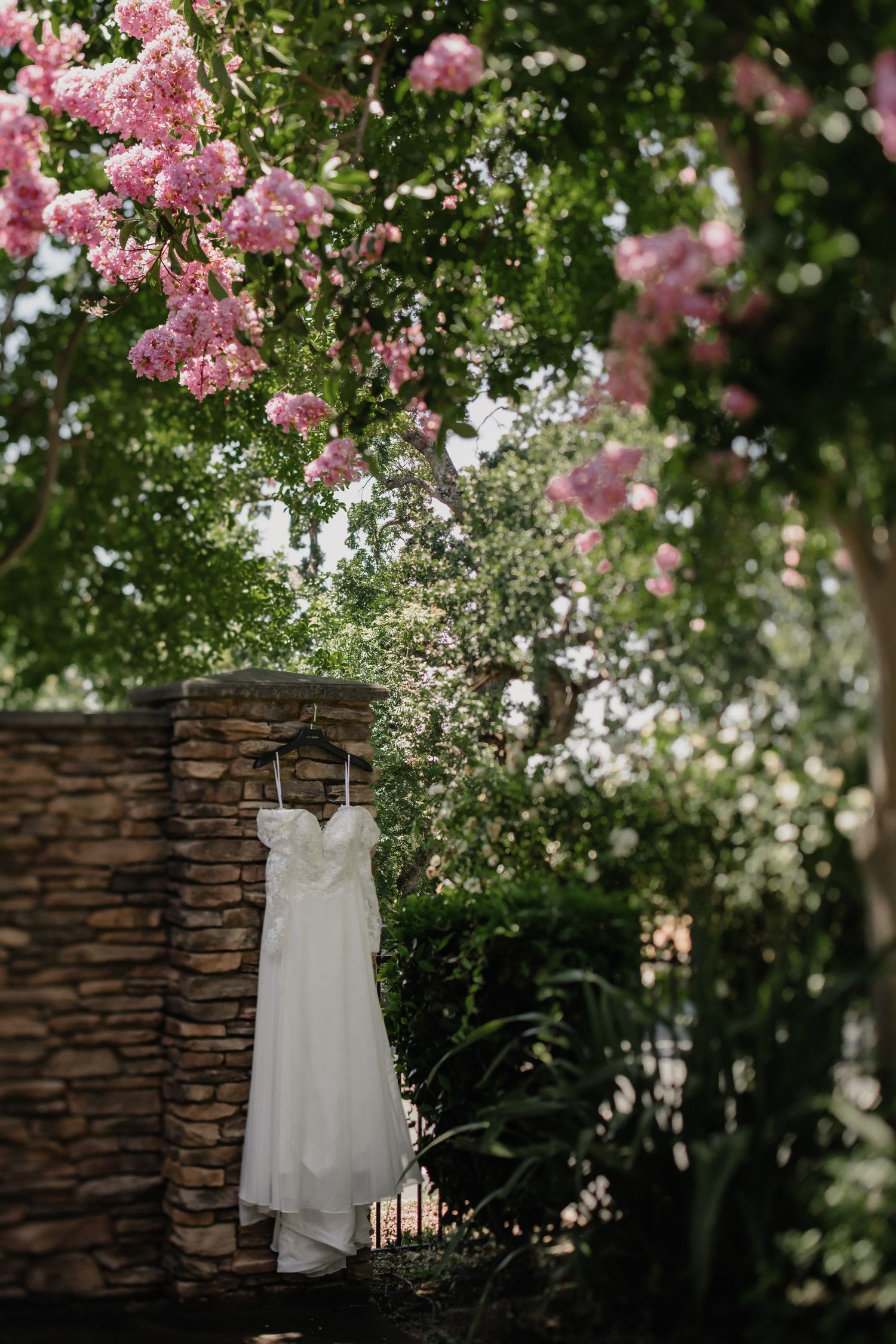 A white wedding dress hangs outdoors against a stone wall surrounded by lush greenery and pink flowers.