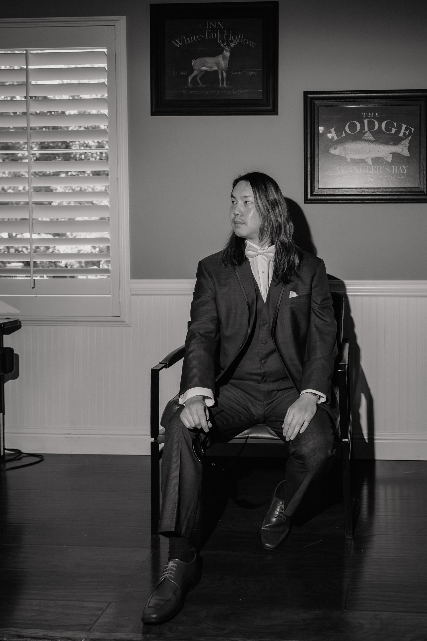 Groom with long hair sits in chair wearing dark suit in black and white wedding portrait.