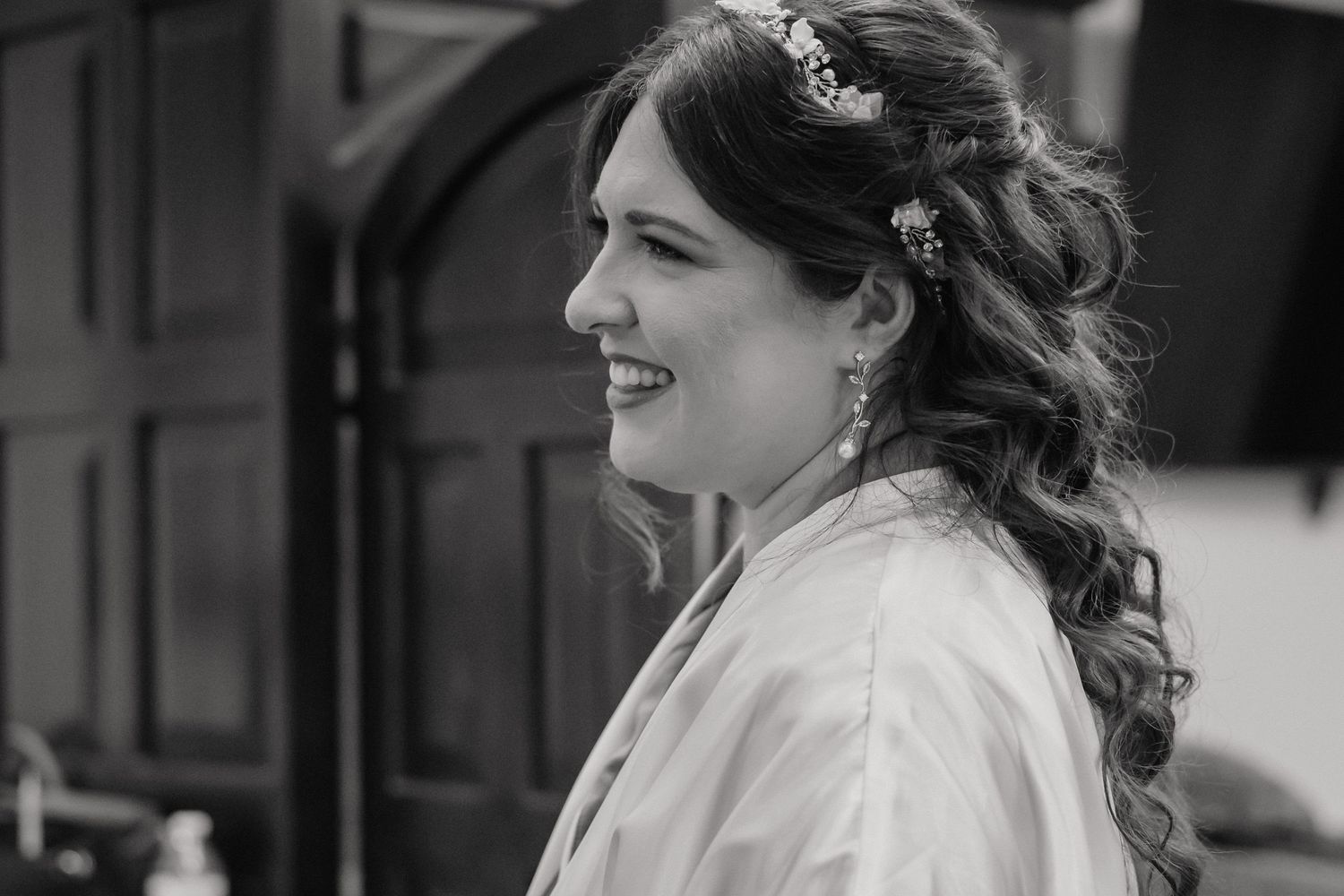 Black and white portrait of a smiling bride with floral hair accessory and elegant earrings.