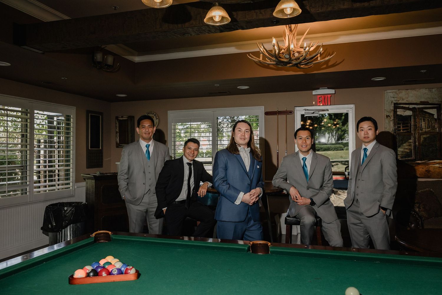Groom and groomsmen pose together in suits beside a pool table before the wedding ceremony.