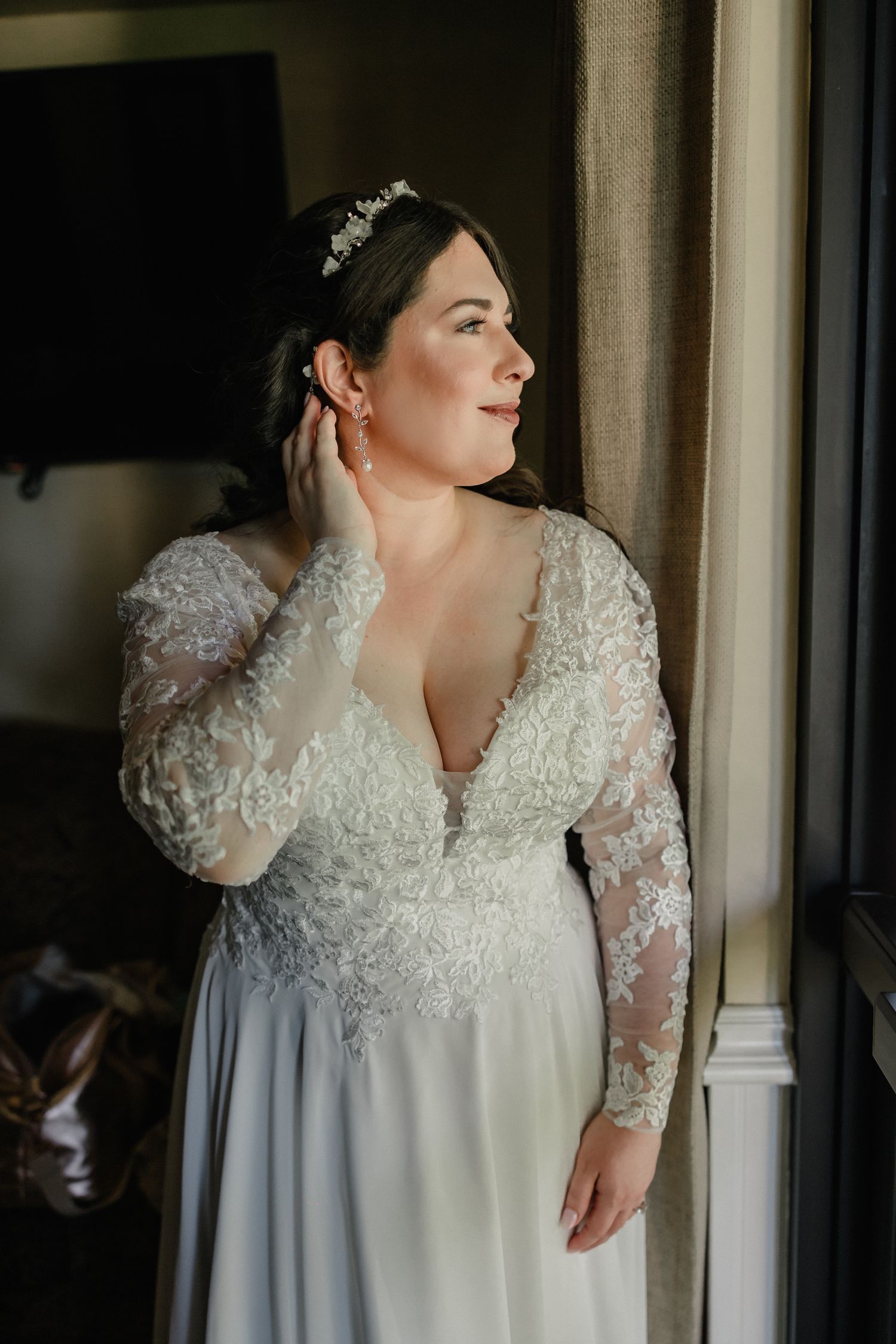 Bride in lace long-sleeve wedding gown with tiara adjusts earring by window.