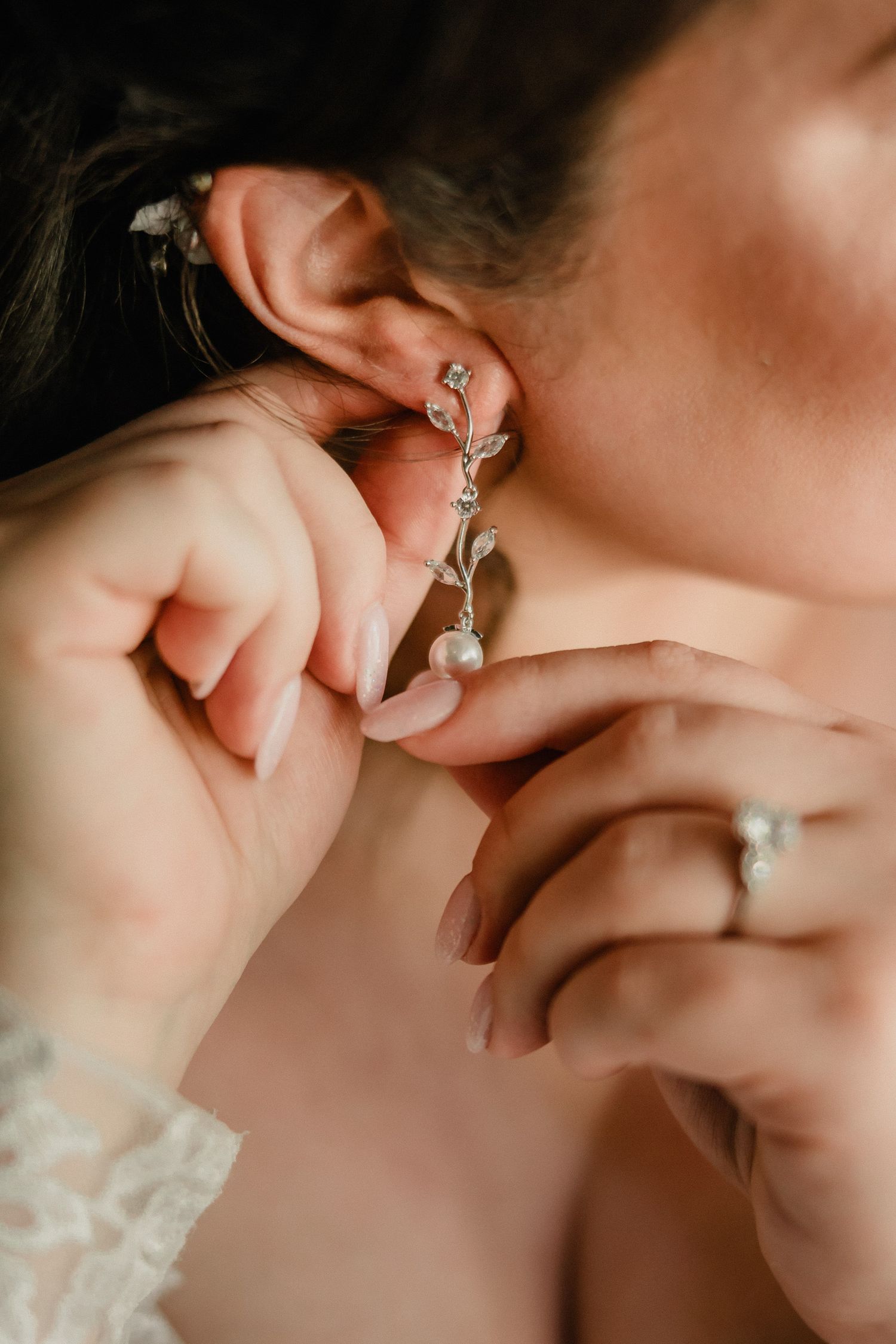 Bride putting on elegant silver leaf and pearl drop earring with diamond engagement ring visible on her finger.