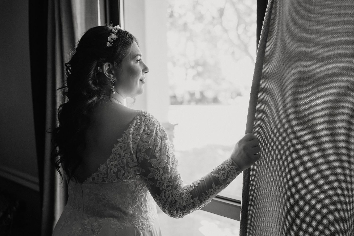 Black and white portrait of bride in lace gown gazing out window.