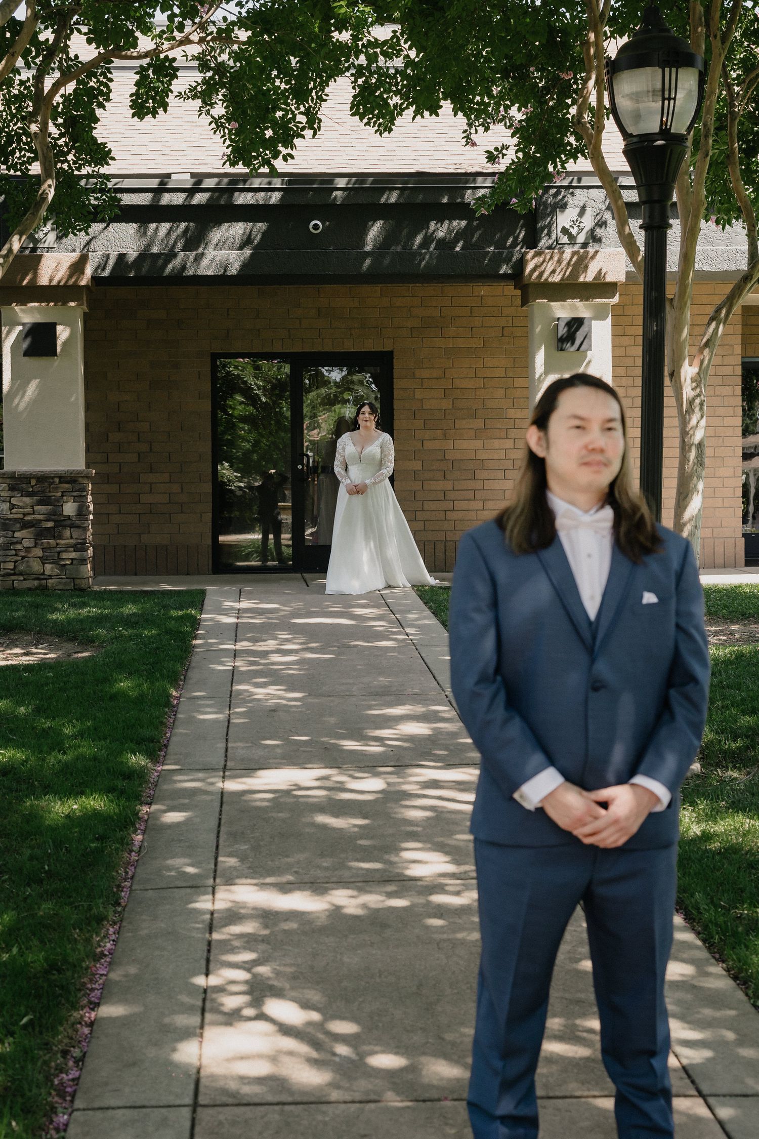 Groom stands on pathway as bride in lace wedding dress approaches for their first look.