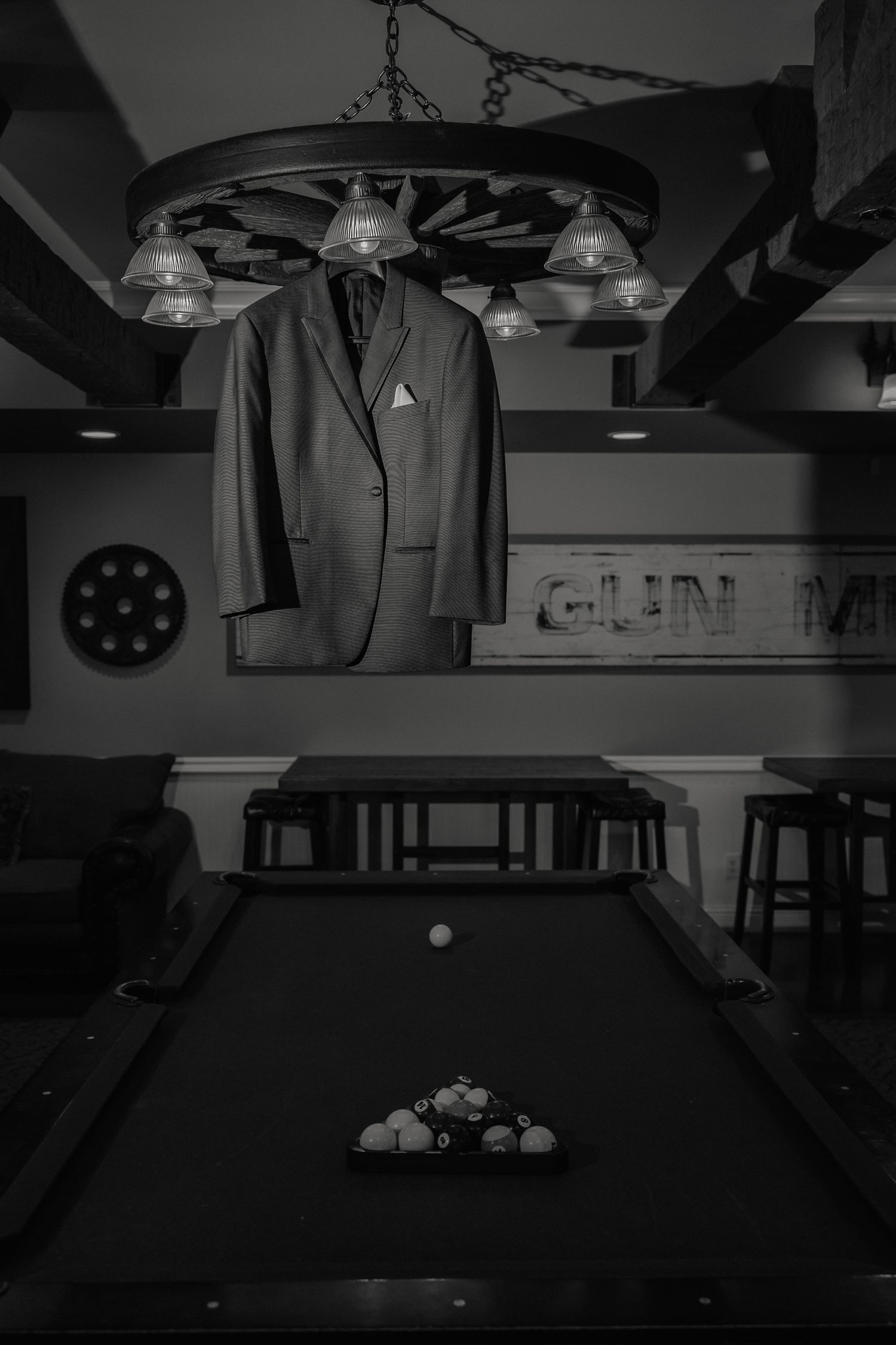 A suit jacket hangs over a pool table beneath a wagon wheel chandelier in a dimly lit game room.