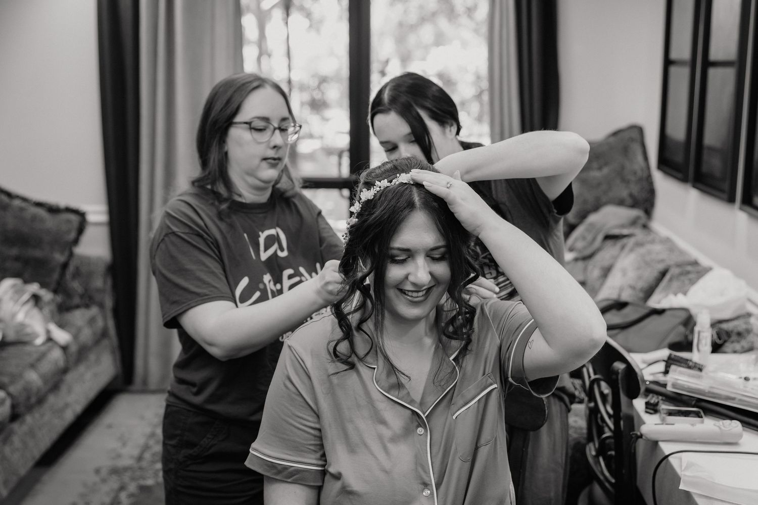 Bridesmaids help smiling bride with tiara placement during black and white wedding preparations.