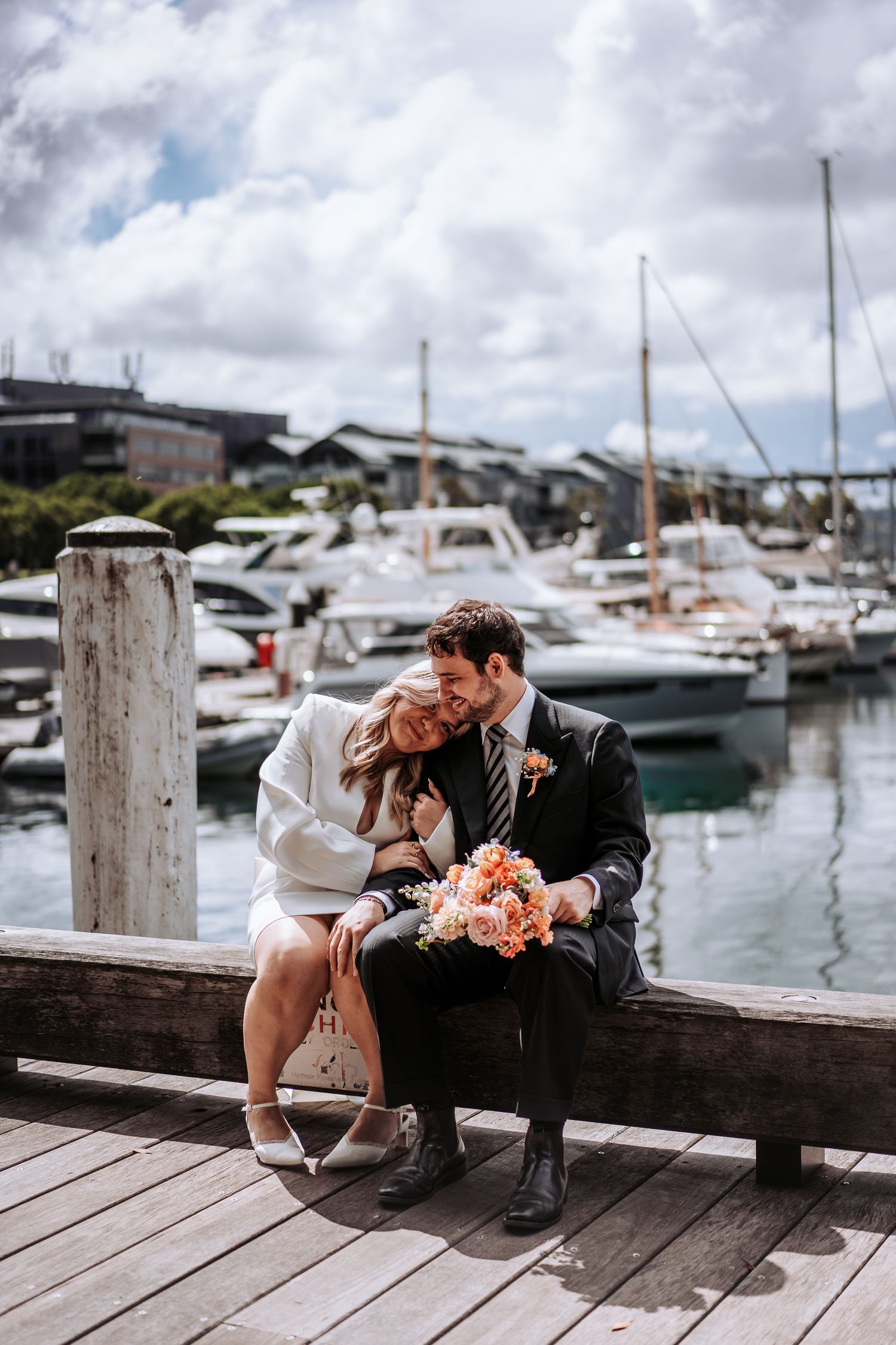 Bride and groom sitting on a wooden dock at a marina, sharing a tender moment with a bridal bouquet.