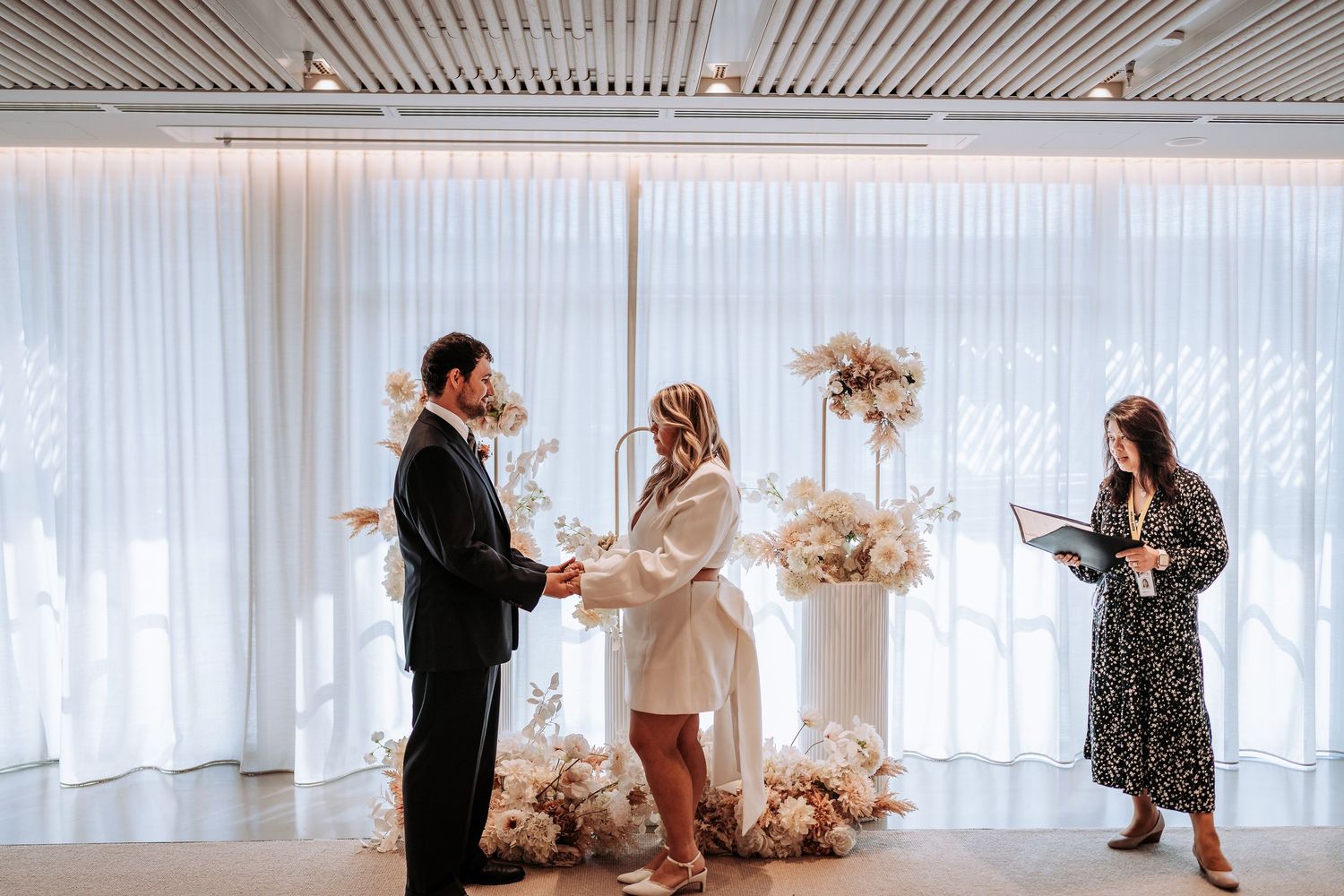Couple exchanging vows at modern wedding ceremony with white floral arrangements and draped backdrop.