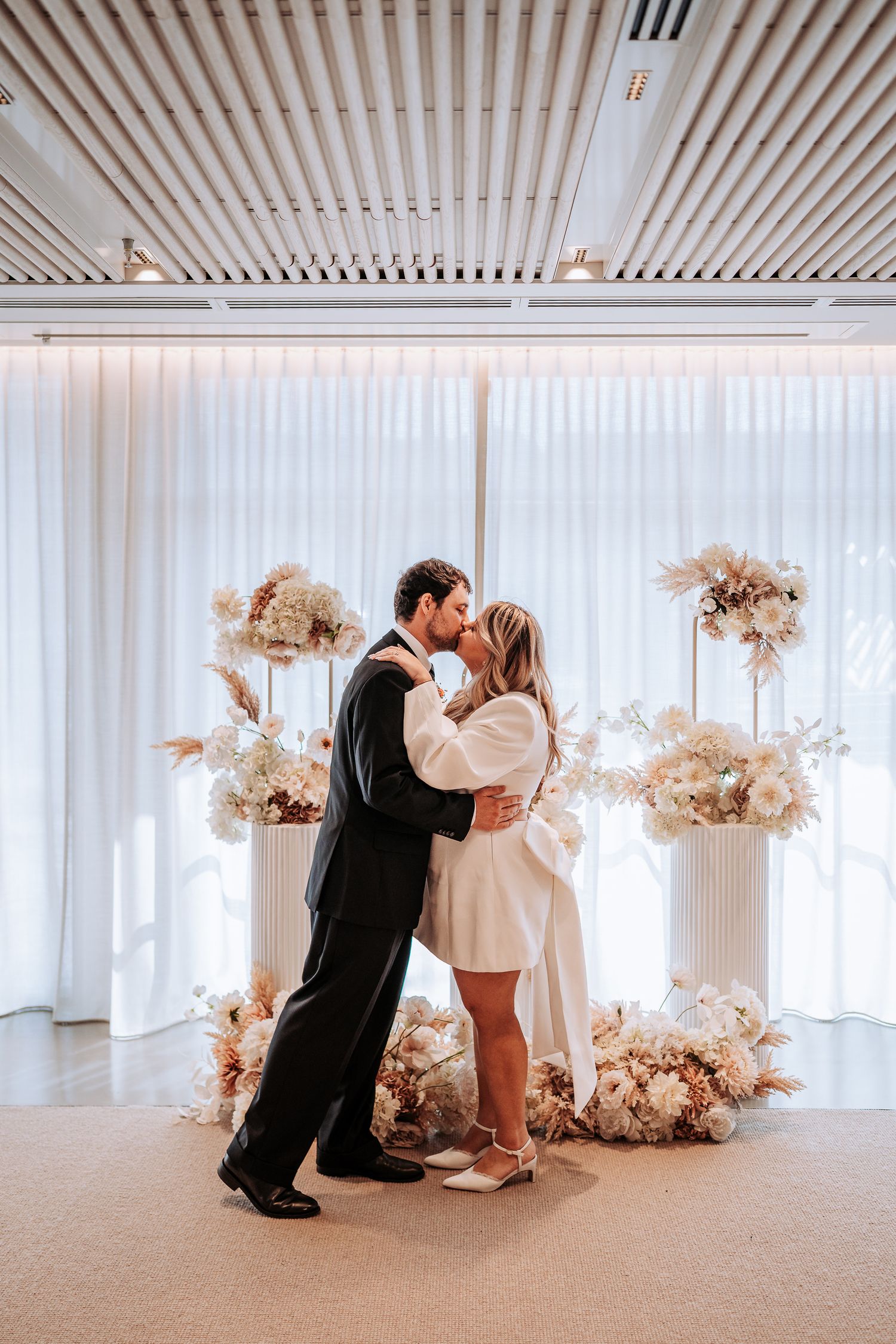 Couple sharing first kiss at wedding altar surrounded by elegant white and blush floral arrangements in modern venue.