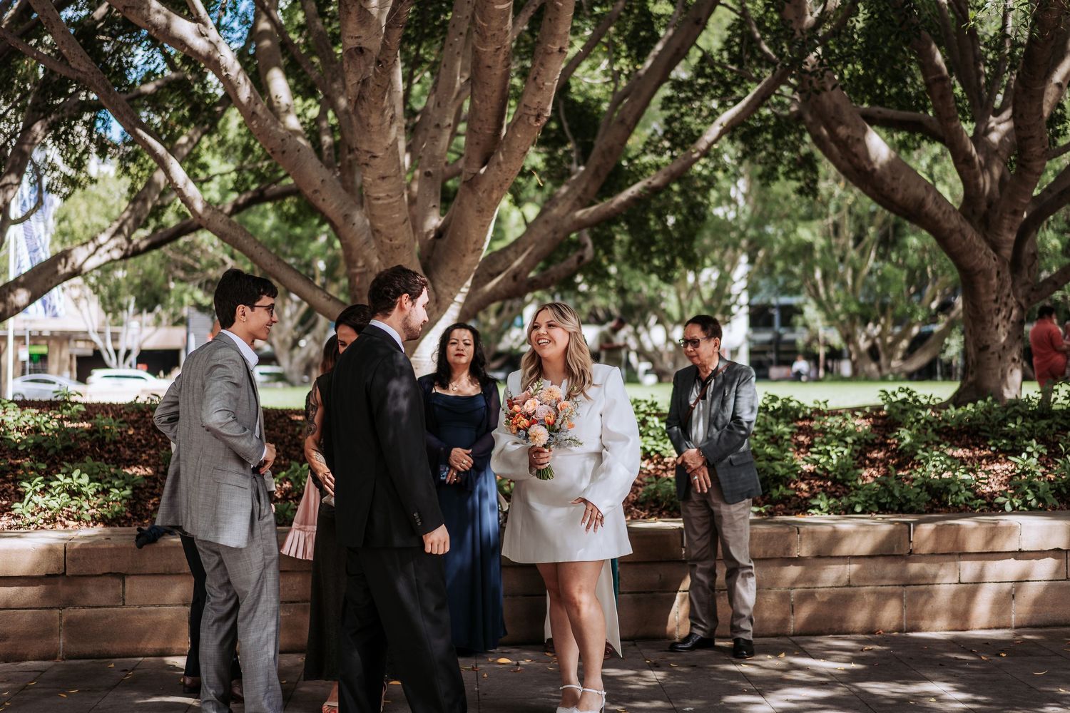 Small wedding group celebrating outdoors under large shady trees in a city park.