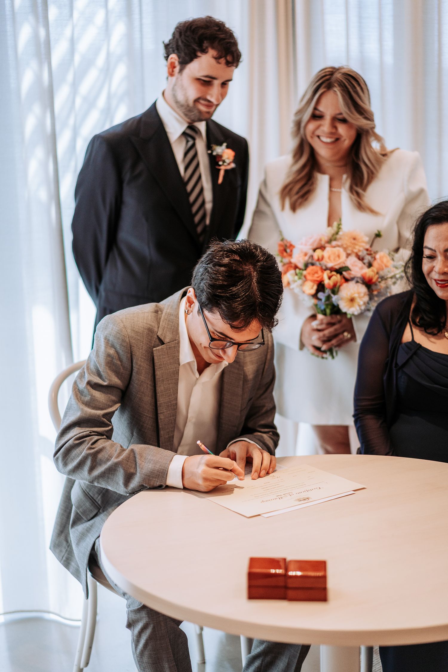 Groom signing wedding certificate at round table while bride and guests look on at intimate ceremony.
