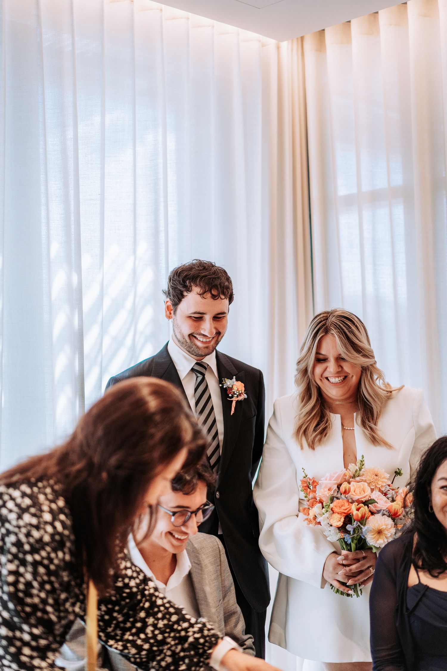 Smiling bride holding peach bouquet and groom in suit signing wedding documents at civil ceremony.
