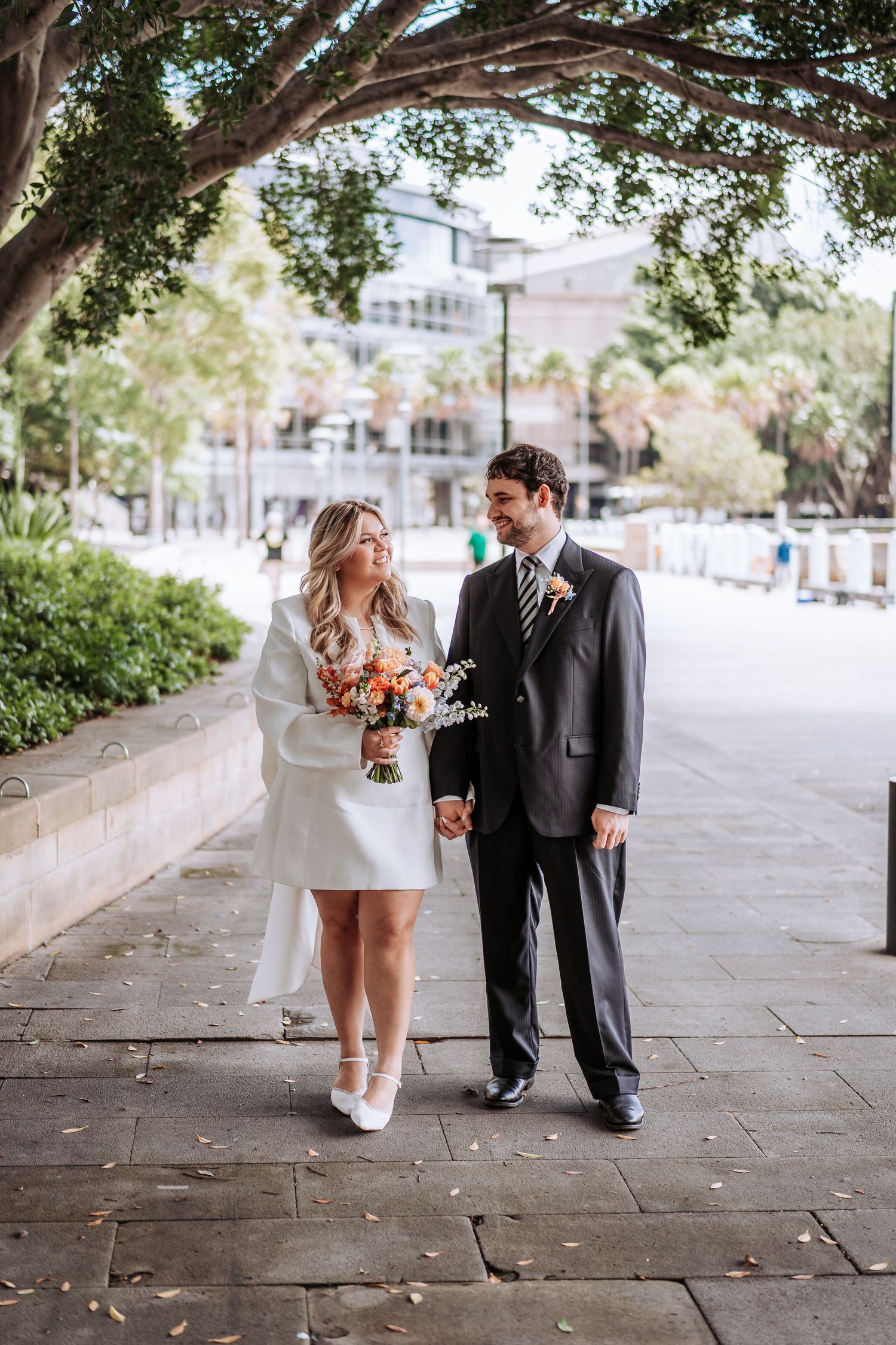 Bride holding orange bouquet and groom in grey suit walk hand in hand along shaded garden pathway.