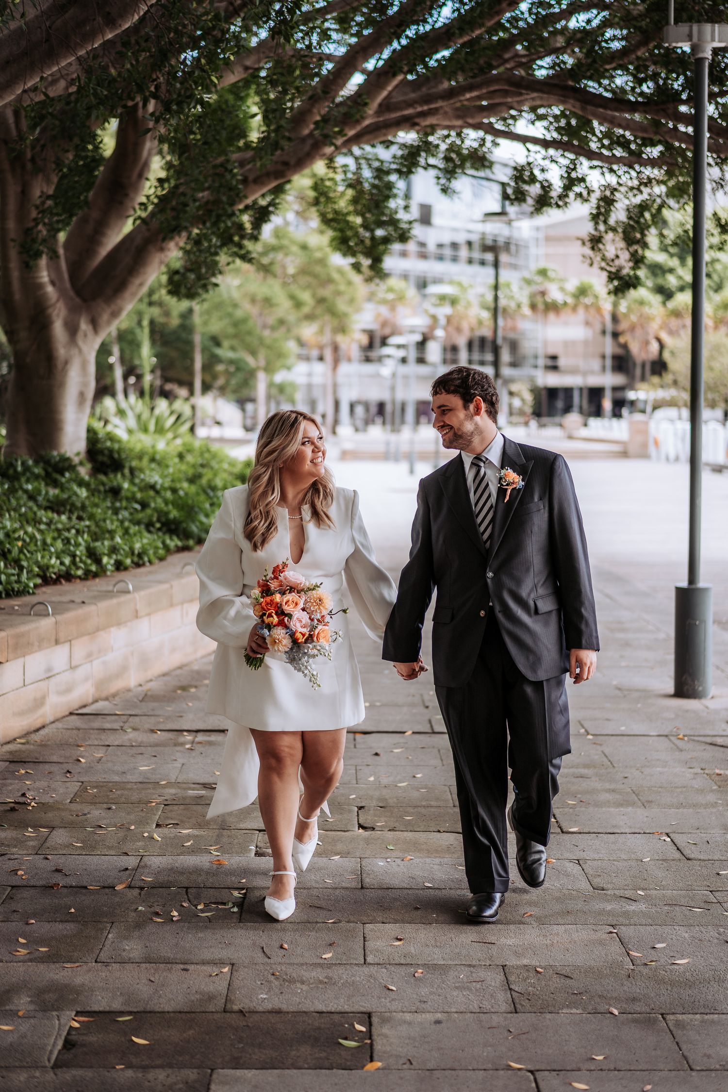 Bride with orange bouquet and groom walking hand in hand along a tree-lined path.