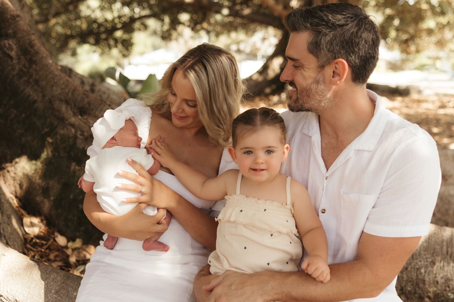 A happy family of four poses outdoors under a tree, with parents holding a newborn and toddler in white outfits.