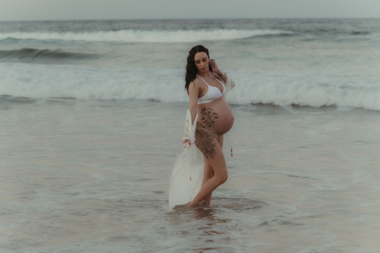 Pregnant woman in white bikini and sheer skirt standing in ocean waves during maternity photoshoot on beach.