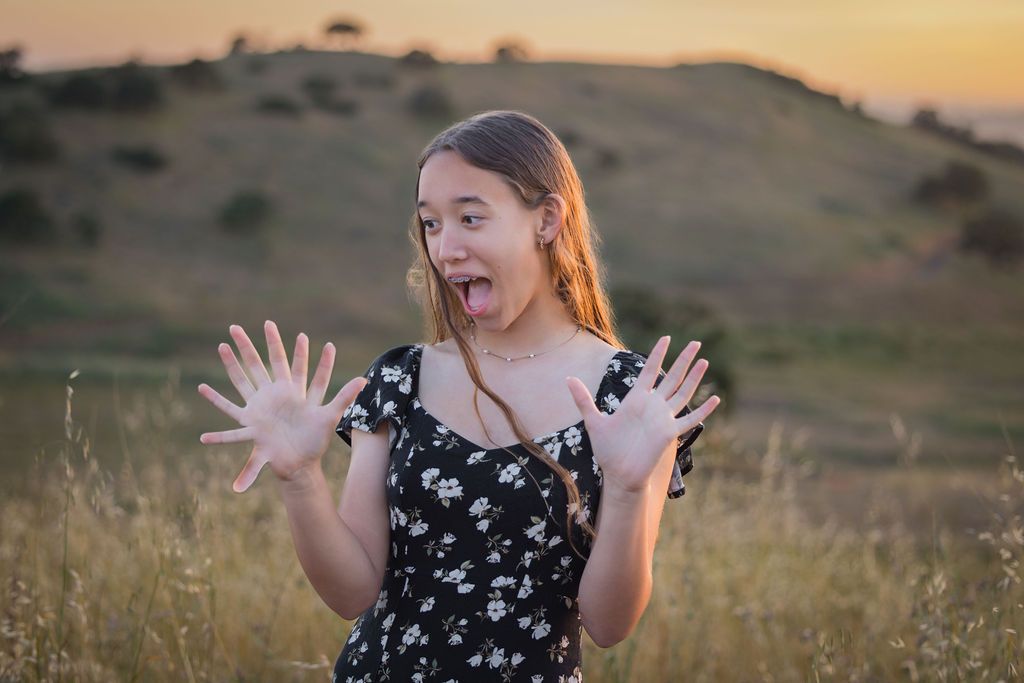 Young woman in floral dress making a surprised face with hands raised outdoors at sunset.