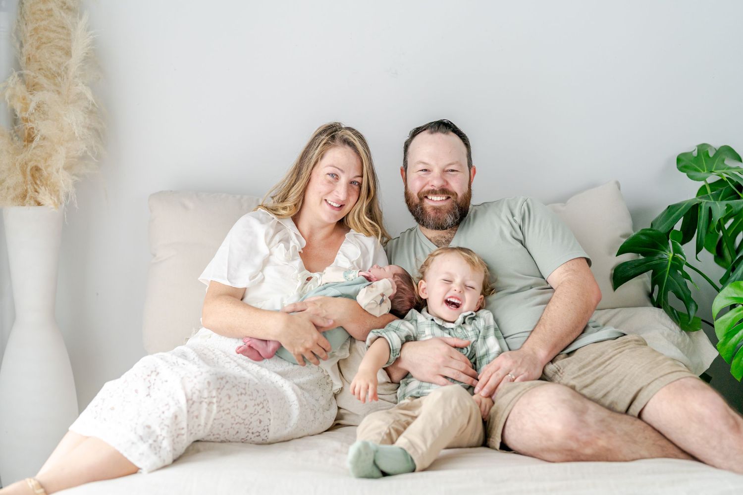 Happy family of four poses on a cozy bed, mom holding newborn baby while toddler laughs with bearded dad.