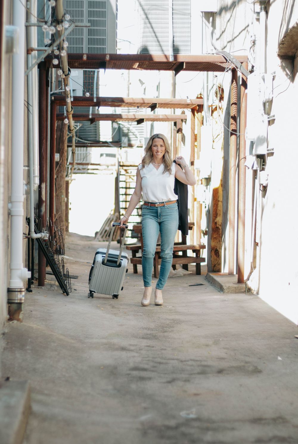Woman in jeans and white blouse pulling silver luggage through an urban alleyway.