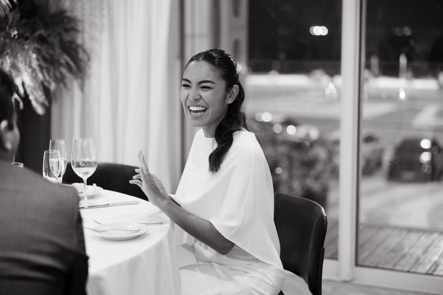 Black and white photo of a laughing woman in white dress seated at a restaurant dinner table.