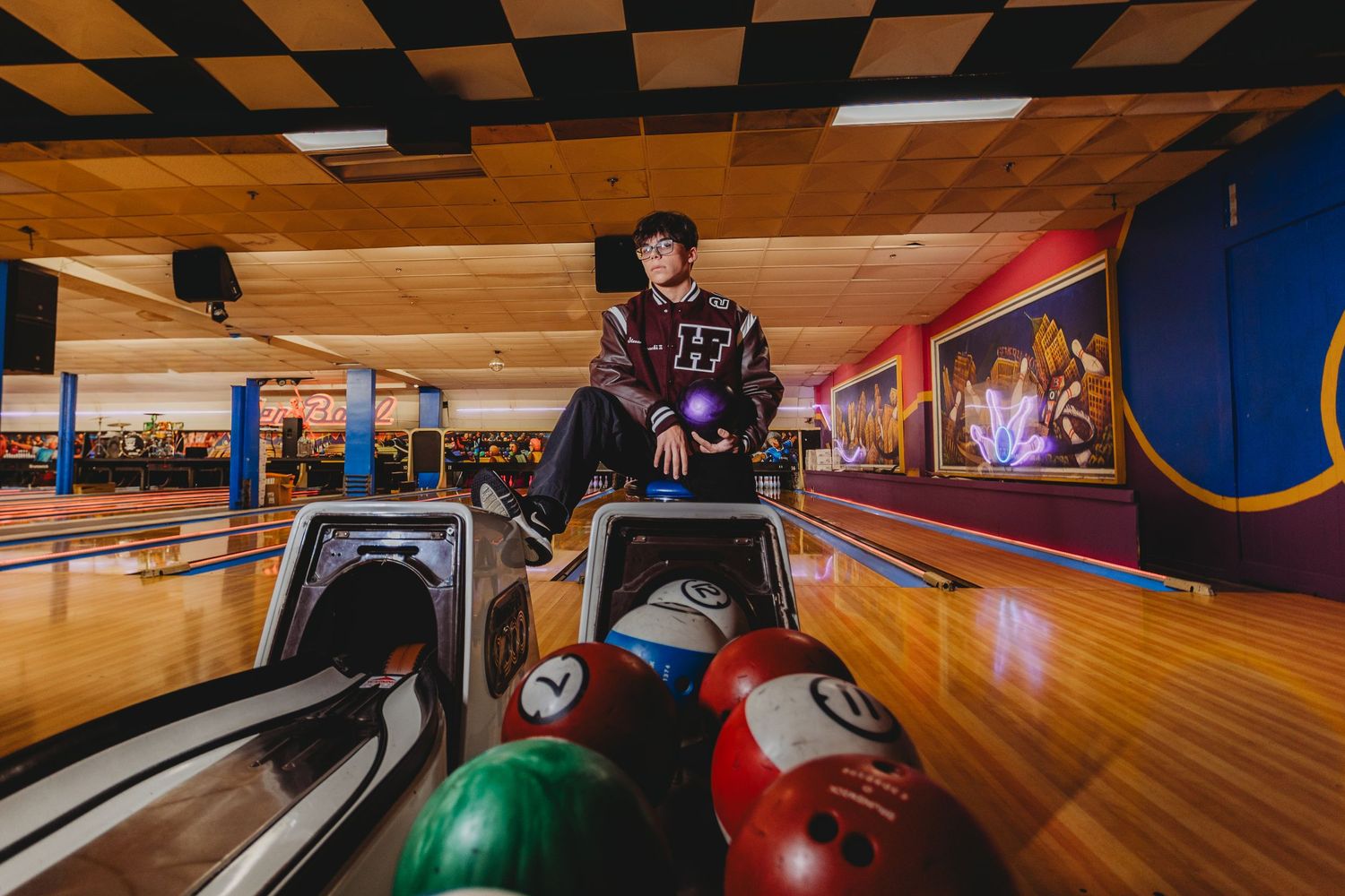 A young man holds a purple bowling ball at a colorful bowling alley, surrounded by multicolored balls on the ball return. detroit senior