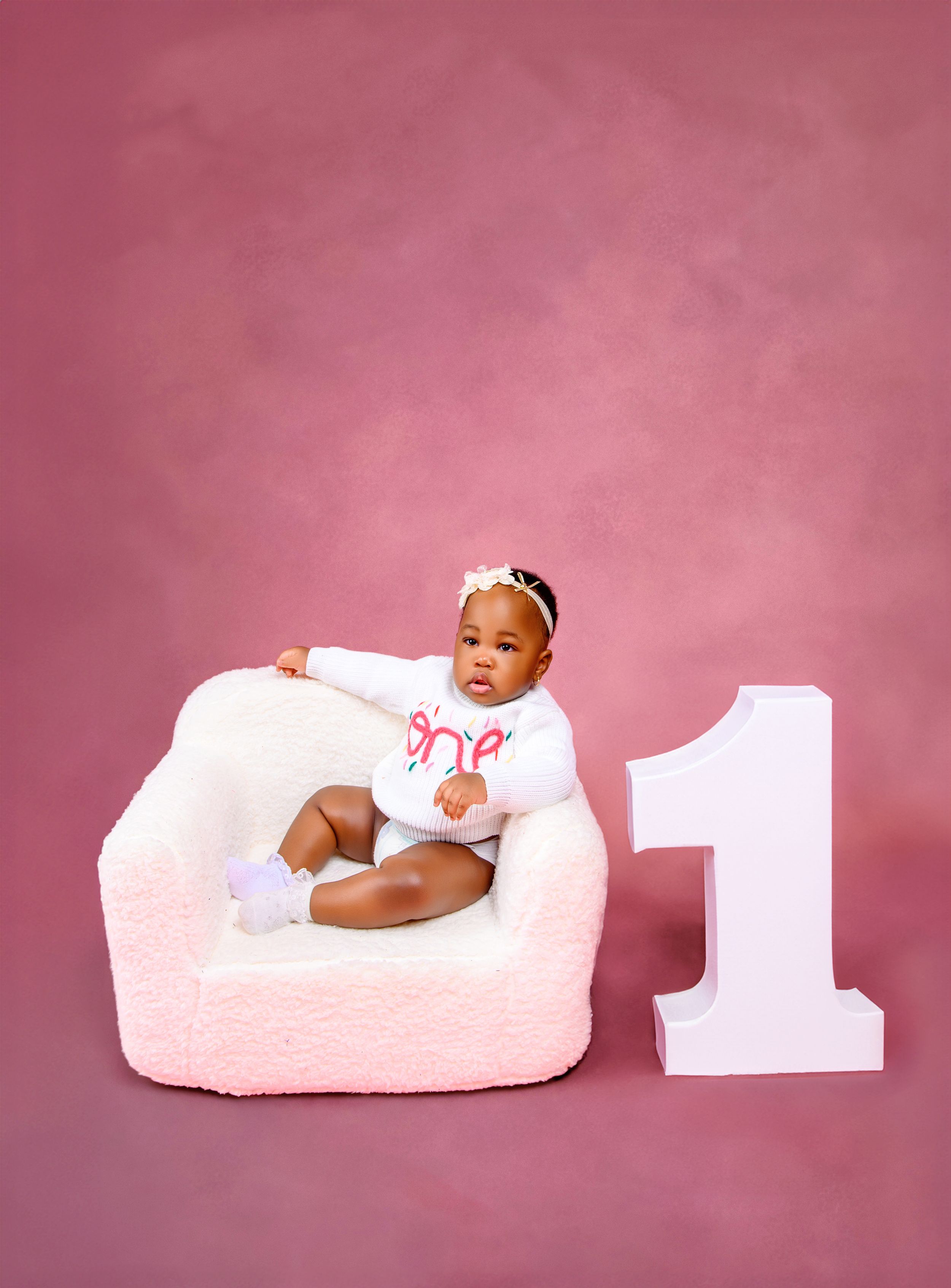 Baby girl in white outfit sits in fluffy pink chair beside a large number 1 on pink backdrop for first birthday photo.