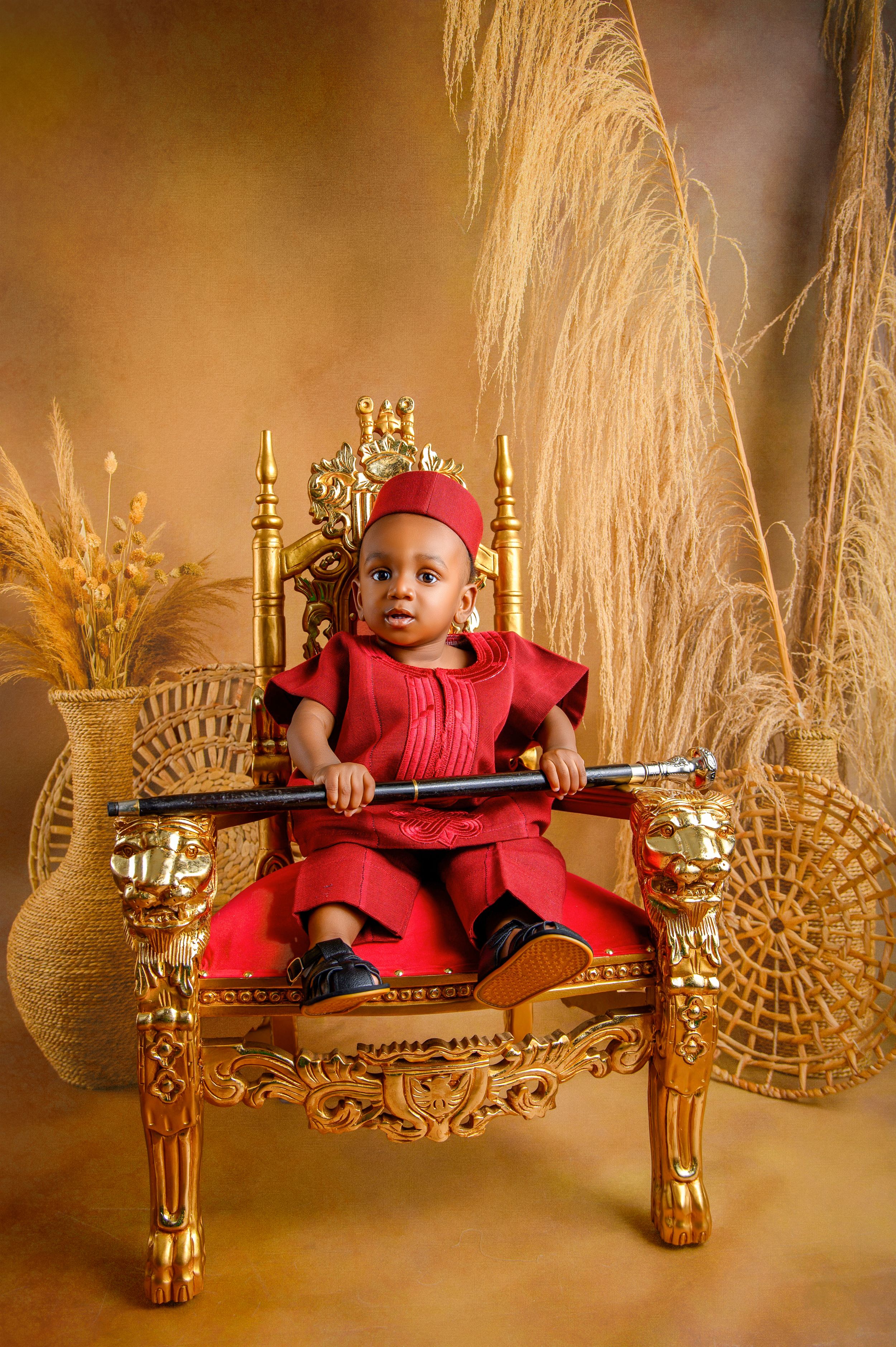Adorable baby boy in red traditional outfit seated on ornate gold throne chair holding a scepter.