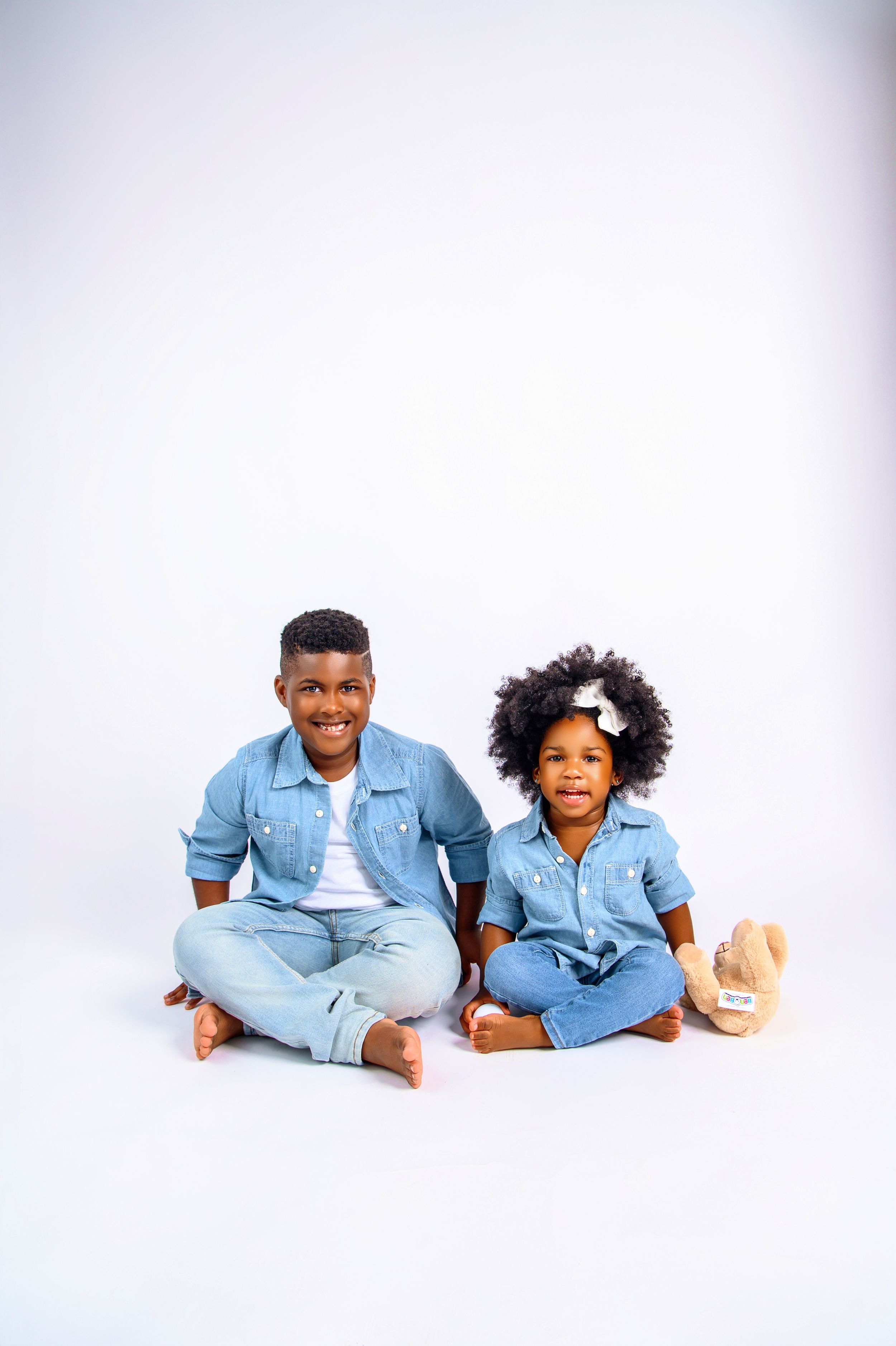 Two smiling Black children in matching denim outfits sit together on a white background, a boy and a curly-haired girl.