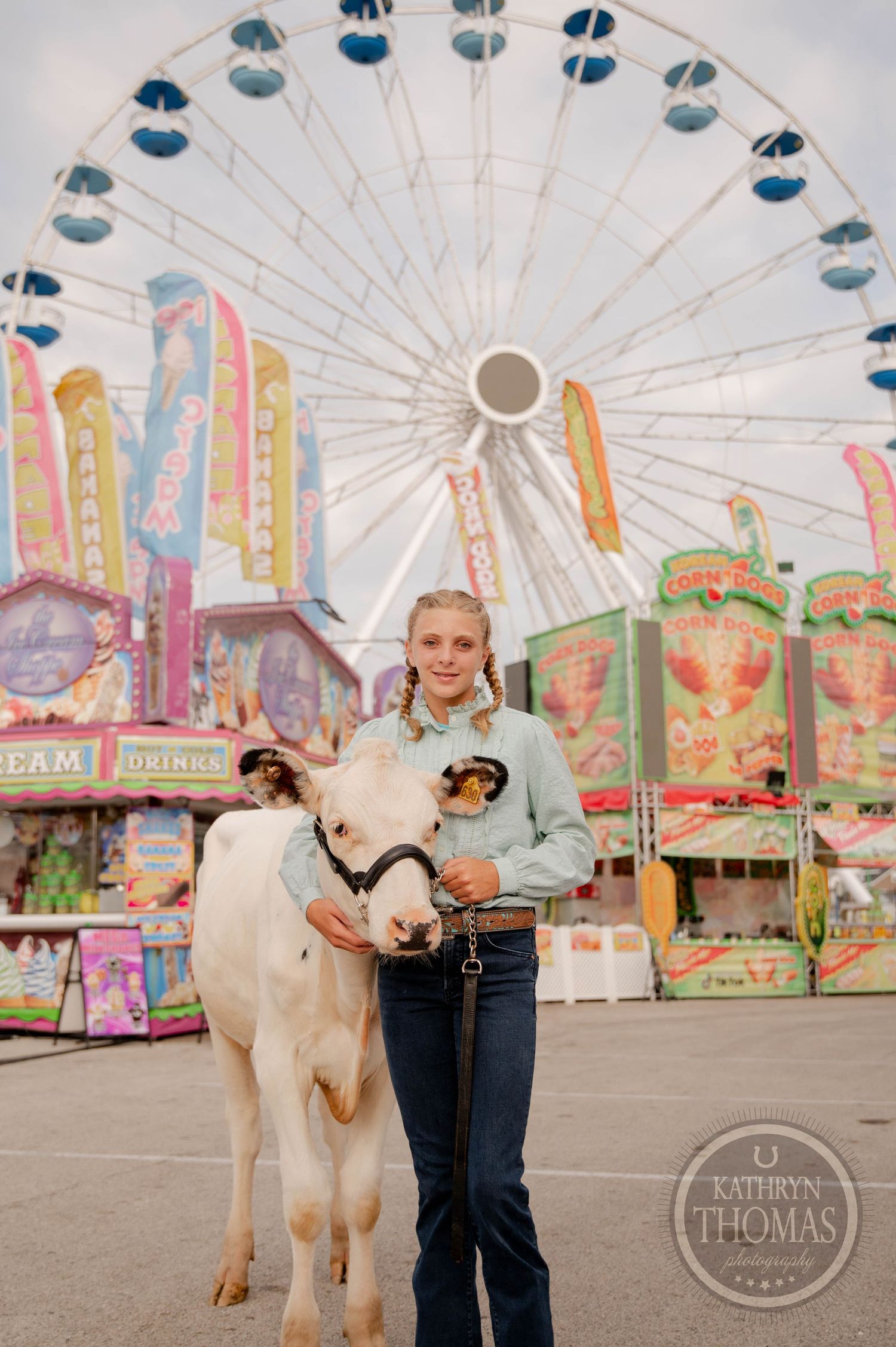 Young girl holding a white calf at a county fair with a Ferris wheel and colorful game booths in the background.