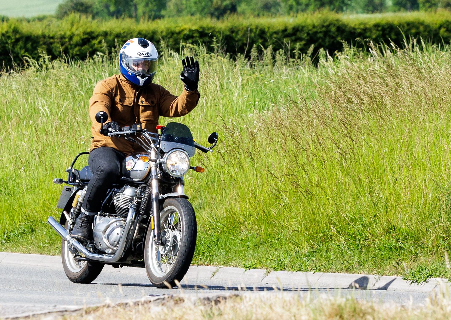 A motorcyclist in a brown jacket and blue helmet waves while riding a classic motorcycle on a rural road.