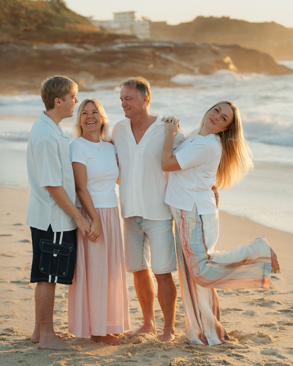 Family of four posing together on a sandy beach at golden hour, wearing coordinated white and pastel outfits at Bronte Beach photoshoot