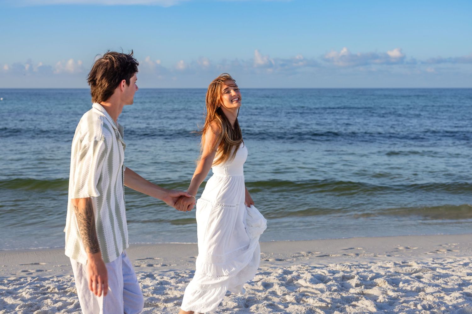 A romantic couple holds hands on a sunny white sand beach with calm blue ocean waves in the background.