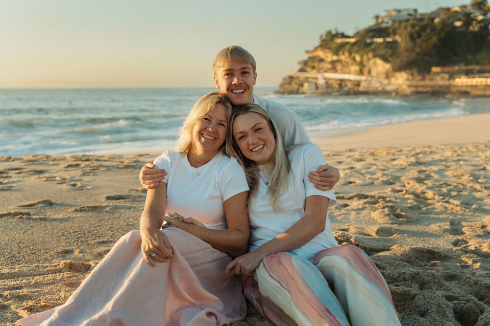 Mom, son, daughter smiling and sit together on a sandy beach at golden photoshoot hour with ocean waves and cliffs in the background Bronte