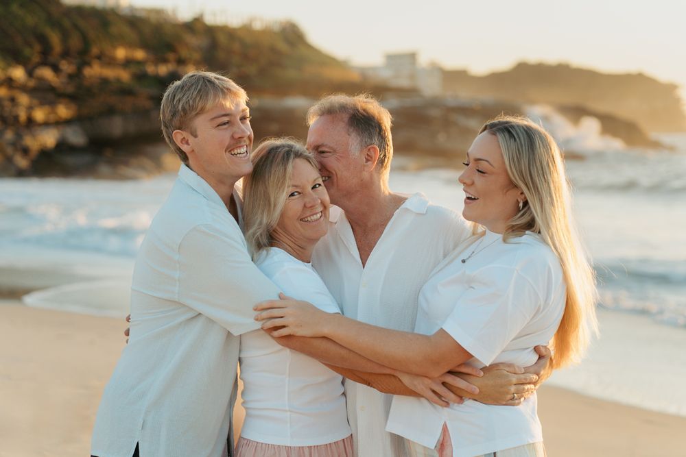 Family of four in white outfits laughing and embracing on a sunny beach at golden hour photoshoot in Sydney Bronte Beach