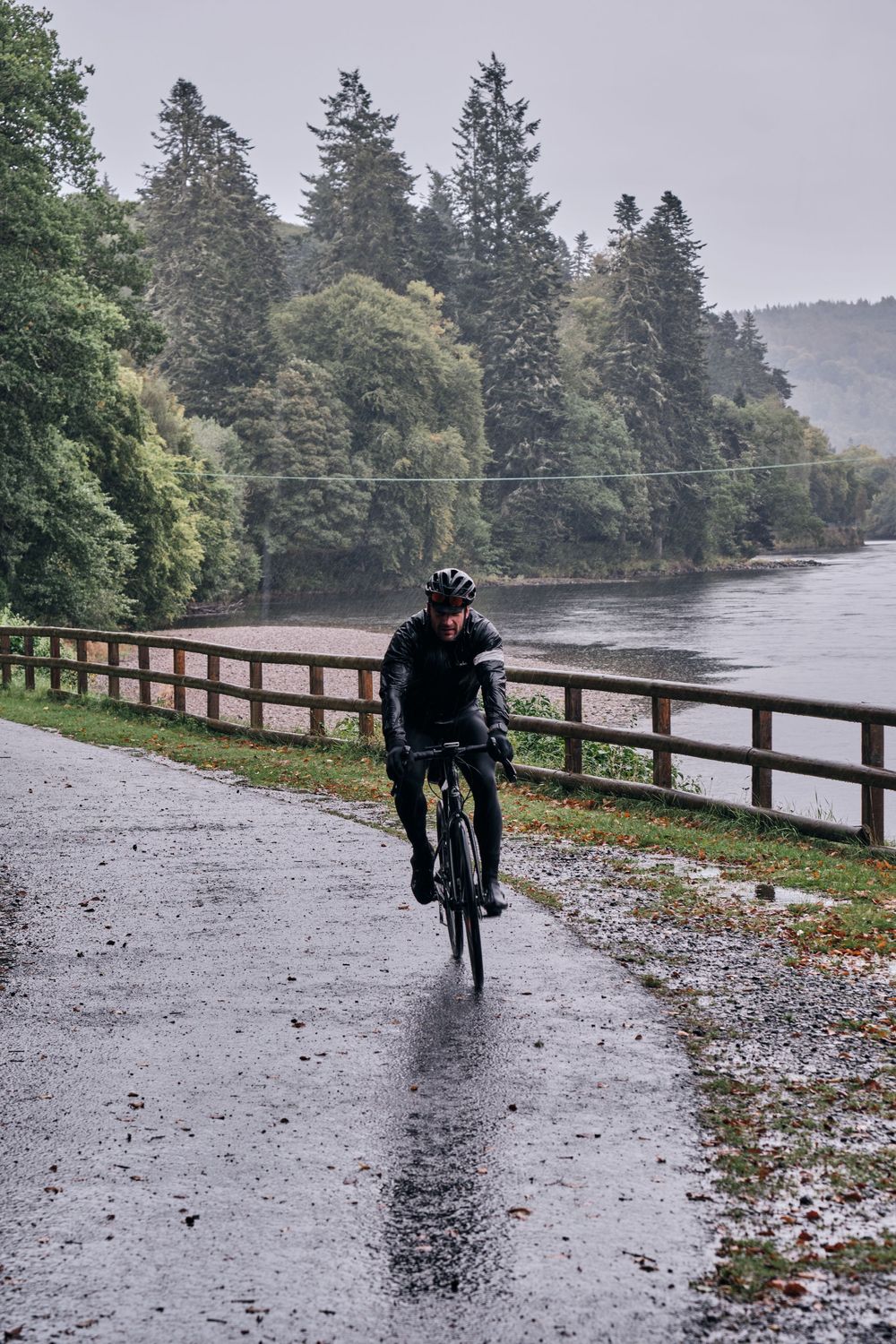 Cyclist riding in heavy rain on a wet road beside a wooden fence and river, surrounded by lush green trees.