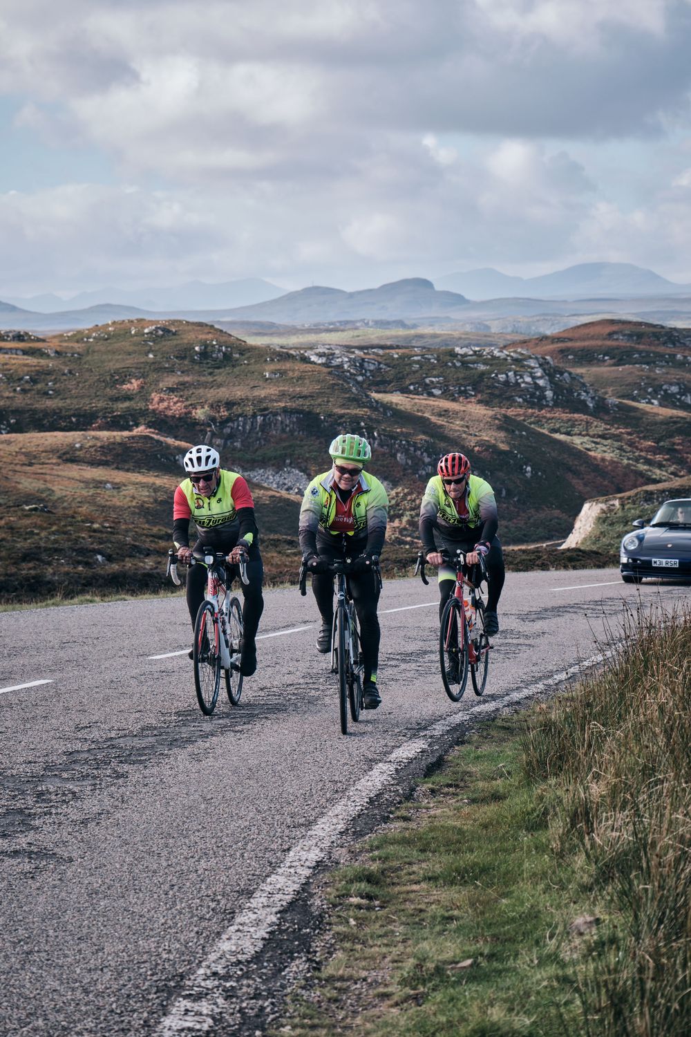 Three cyclists in bright gear ride road bikes through scenic Scottish Highlands with mountains in the background.