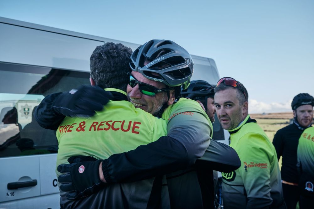 Cyclists in neon yellow jackets share an emotional hug with a rescue worker outdoors near a white van.